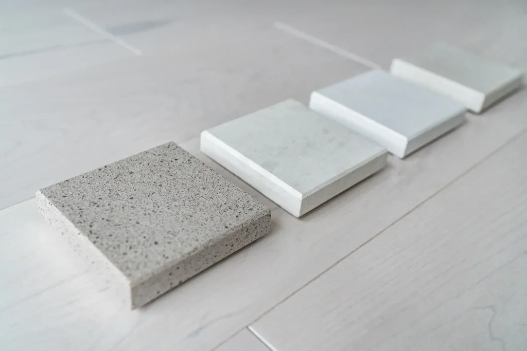 Vier quadratische Fliesenmuster in verschiedenen Grau- und Beigetönen liegen in einer Reihe auf einem hellen Holzboden, um die Wirkung der Materialien und Texturen im realen Wohnumfeld zu vergleichen.