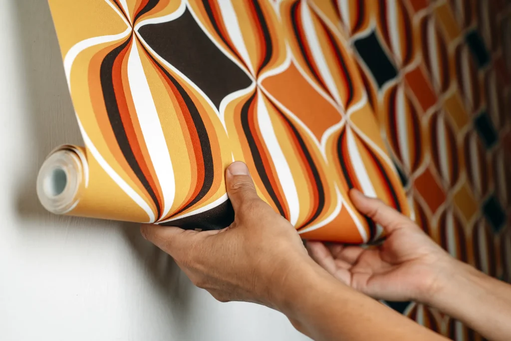Eine Nahaufnahme zeigt eine Person, die eine bunte Mustertapete mit Retro-Design in Orange-, Gelb-, Braun- und Weißtönen an eine Wand anbringt.