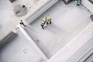 Luftaufnahme einer Baustelle mit Bauingenieuren und Arbeitern in Sicherheitswesten und Helmen, die eine Betonkonstruktion inspizieren. Sichtbar sind Betonwände und eine gegossene Betonfläche, die verschiedene Betonarten im Bauprozess veranschaulichen.