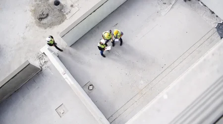 Luftaufnahme einer Baustelle mit Bauingenieuren und Arbeitern in Sicherheitswesten und Helmen, die eine Betonkonstruktion inspizieren. Sichtbar sind Betonwände und eine gegossene Betonfläche, die verschiedene Betonarten im Bauprozess veranschaulichen.