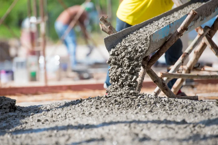 Frisch gemischter Beton fließt aus einer Rutsche auf den Boden einer Baustelle. Im Hintergrund sind Betonbaur in Schutzkleidung zu sehen, die mit Bauarbeiten beschäftigt sind.