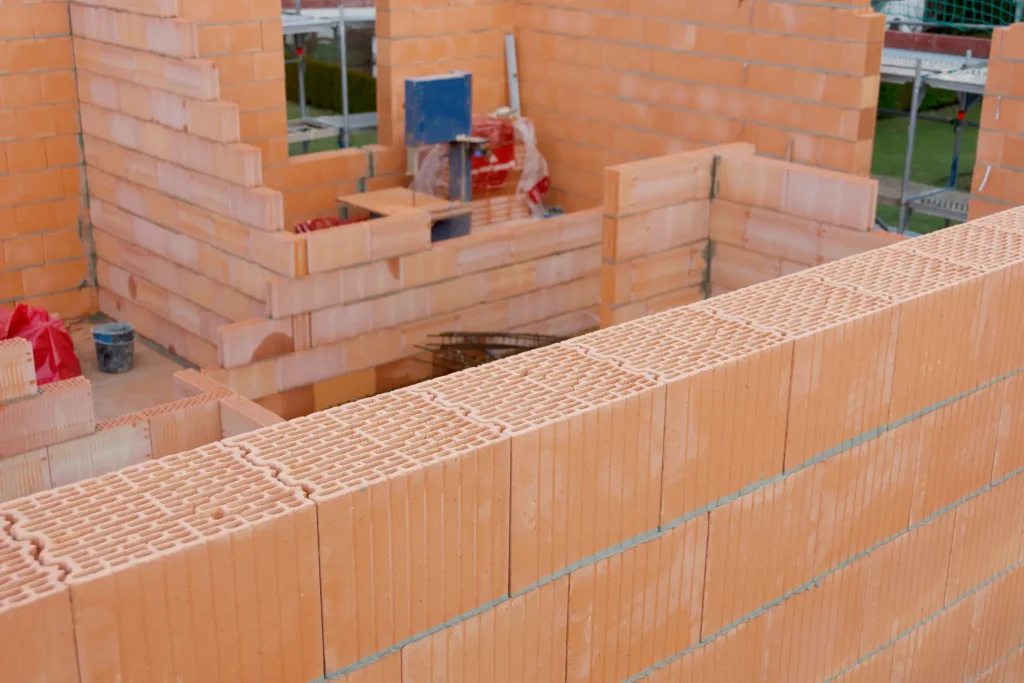 Rohbau eines Einfamilienhauses mit Wänden aus roten Hochlochziegeln. Die Ziegelmauern sind im Baufortschritt sichtbar, mit frischem Mörtel zwischen den Steinen. Baustellenmaterialien und Werkzeuge befinden sich im Hintergrund.