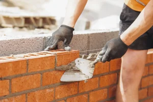 Maurer beim Bau einer Ziegelmauer auf einer Baustelle. Er trägt schwarze Arbeitshandschuhe und verwendet eine Maurerkelle, um Mörtel zwischen die roten Ziegelsteine aufzutragen. Eine Richtschnur sorgt für eine gerade Ausrichtung der Mauer.