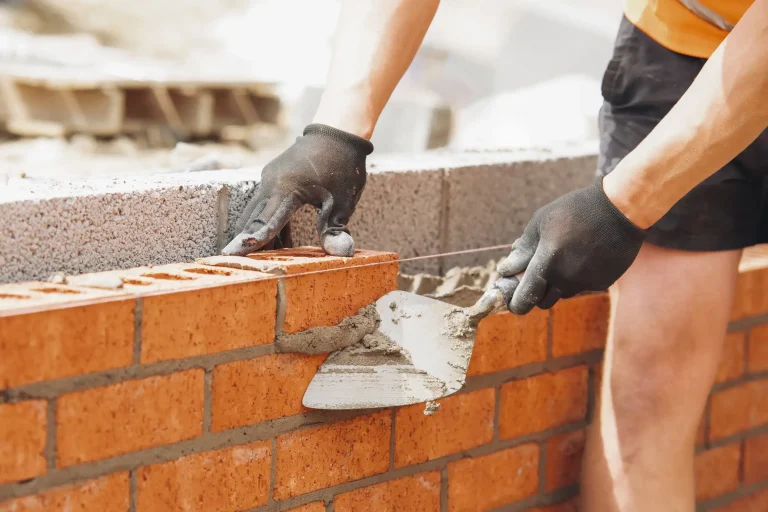 Maurer beim Bau einer Ziegelmauer auf einer Baustelle. Er trägt schwarze Arbeitshandschuhe und verwendet eine Maurerkelle, um Mörtel zwischen die roten Ziegelsteine aufzutragen. Eine Richtschnur sorgt für eine gerade Ausrichtung der Mauer.