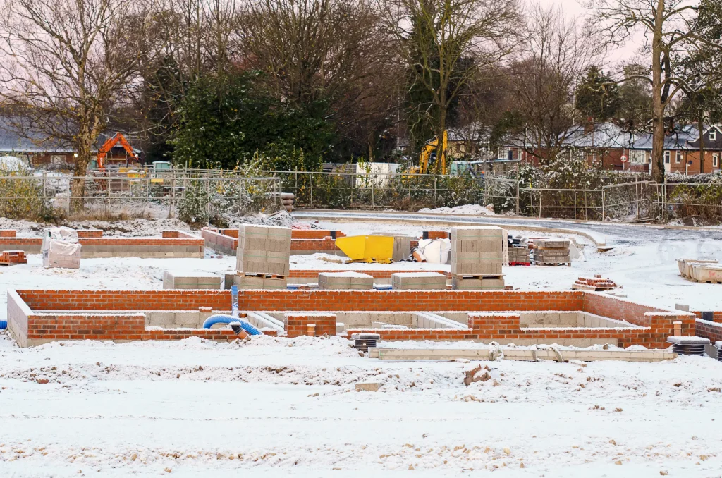 Eine verschneite Baustelle mit teilweise errichteten Fundamenten aus roten Ziegeln und Betonblöcken. Auf dem Gelände liegen Baumaterialien wie Betonsteine, Rohre und ein gelber Bauschubkarren. Im Hintergrund sind Gerüste, Bäume ohne Laub und einige Häuser zu sehen. Der Boden und die Baumaterialien sind mit einer dünnen Schneeschicht bedeckt, was auf winterliche Bedingungen hinweist.