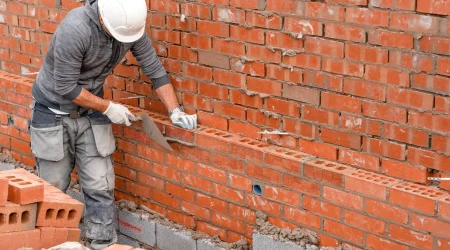 Ein Bauarbeiter mit weißem Schutzhelm und grauem Hoodie mauert eine Wand aus roten Lochziegeln. Er trägt Arbeitshandschuhe und verwendet eine Kelle, um Mörtel auf die Ziegel aufzutragen. Die Ziegelwand ist teilweise fertiggestellt, darunter sind Betonsteine sichtbar. Im Vordergrund liegen weitere rote Ziegel bereit.