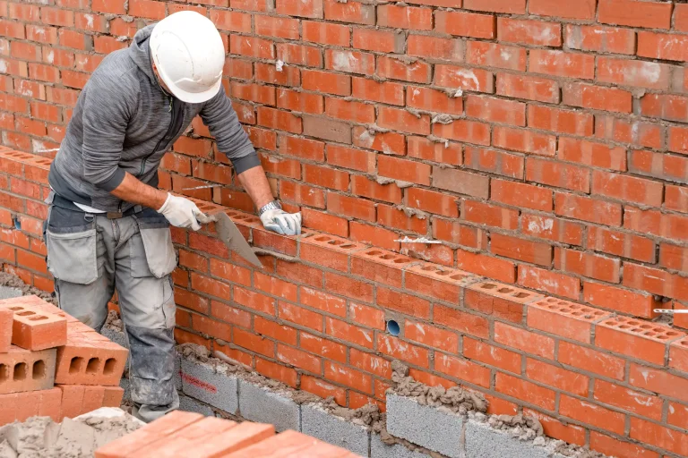Ein Bauarbeiter mit weißem Schutzhelm und grauem Hoodie mauert eine Wand aus roten Lochziegeln. Er trägt Arbeitshandschuhe und verwendet eine Kelle, um Mörtel auf die Ziegel aufzutragen. Die Ziegelwand ist teilweise fertiggestellt, darunter sind Betonsteine sichtbar. Im Vordergrund liegen weitere rote Ziegel bereit.