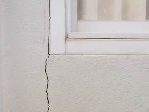 Langer vertikaler Riss in einer weißen, verputzten Betonwand, der sich von unten bis zum Fensterrahmen erstreckt. Das Fenster hat einen weißen Kunststoffrahmen mit einer leicht verschmutzten unteren Kante. Die Glasscheibe ist milchig und reflektiert leicht das Umfeld.