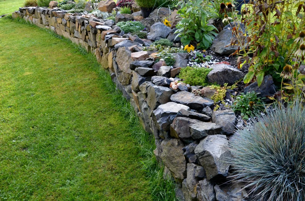 Trockenmauer aus Natursteinen als Gartenbegrenzung mit bepflanztem Hochbeet. Verschiedene Stauden, Gräser und Blühpflanzen wachsen zwischen den Steinen und sorgen für eine naturnahe Gestaltung. Die Mauer stabilisiert das Gelände und fügt sich harmonisch in die Gartenlandschaft ein.