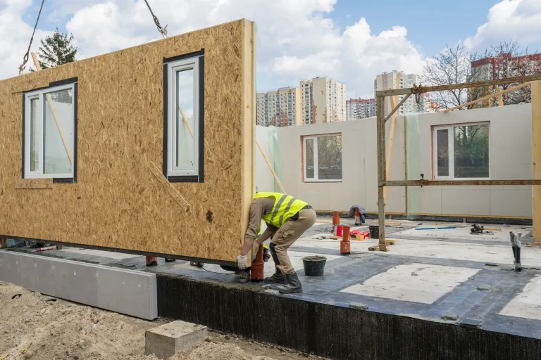 Montage eines vorgefertigten Wandmoduls in Leichtbauweise mit integrierten Fenstern auf einer Baustelle – moderner, nachhaltiger Hausbau in Modul- und Holzrahmenbauweise.