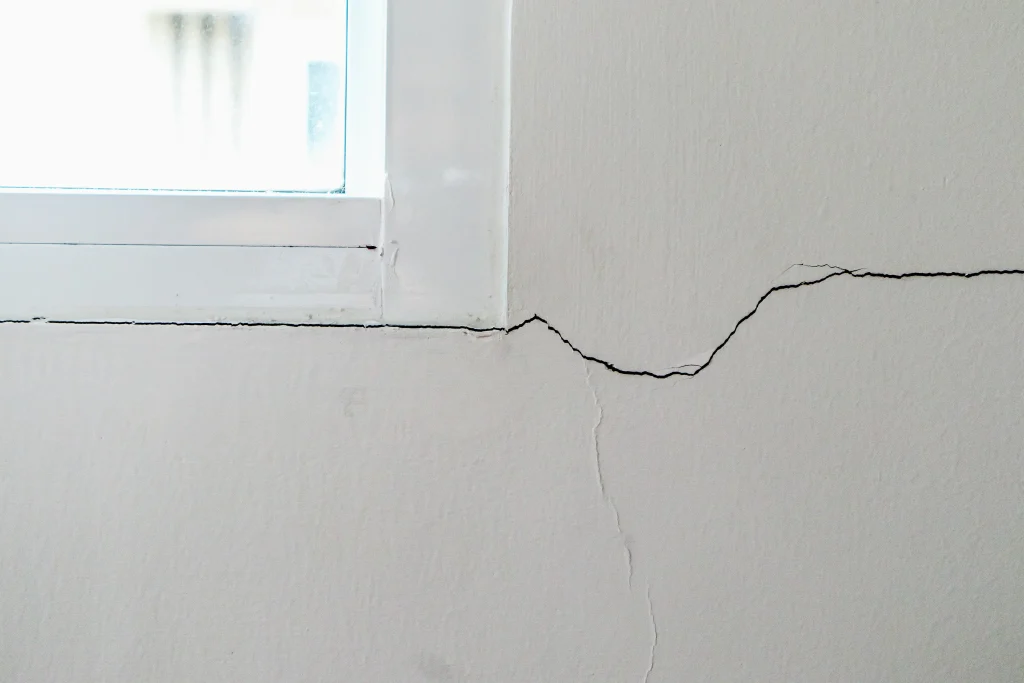 Riss in einer weißen Innenwand unterhalb eines Fensters mit Kunststoffrahmen.