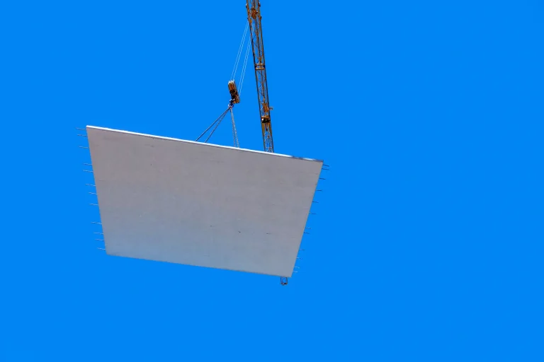 Ein großes Betonfertigteil wird mit Hilfe eines Krans und Stahlseilen in die Luft gehoben. Der Hintergrund zeigt einen klaren, blauen Himmel. An den Seiten des Betonteils sind mehrere herausstehende Bewehrungsstäbe zu sehen.