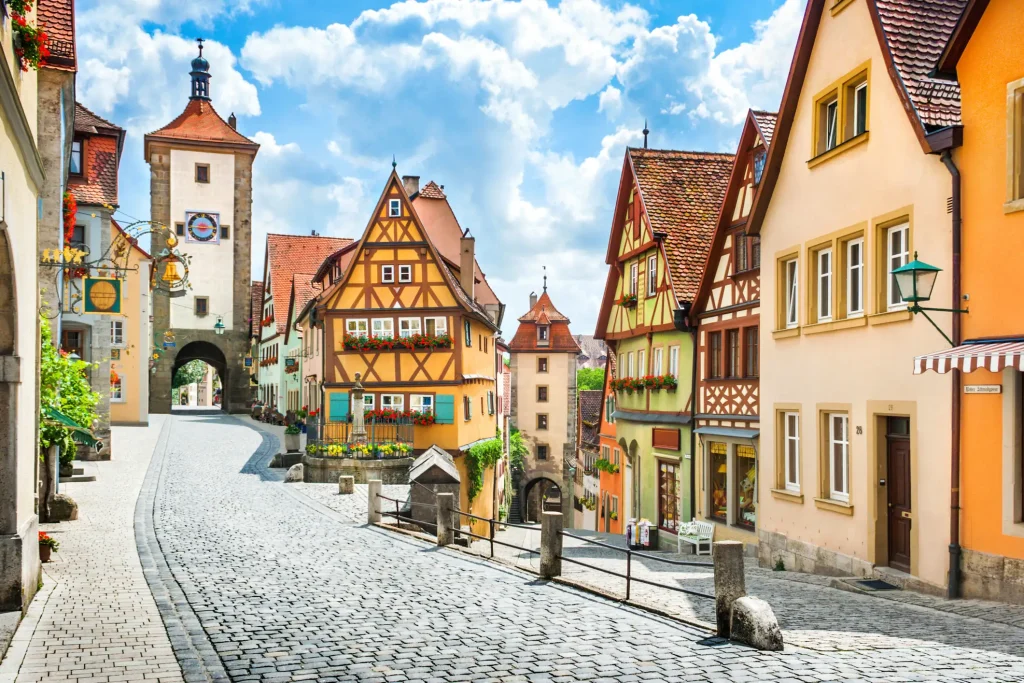 Historisches Stadtbild in Rothenburg ob der Tauber mit Kopfsteinpflasterstraße, mittelalterlichen, denkmalgeschützen Fachwerkhäusern in leuchtenden Farben, Blumen an Fenstern und einem markanten Torturm mit Uhr und Spitzdach im Hintergrund.