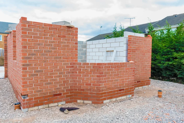 Teilweise errichtete Außenwand eines Neubaus mit rotem Verblendmauerwerk und dahinterliegendem weißen Kalksandstein, im Vordergrund Schotterboden mit Baustellenmaterial.