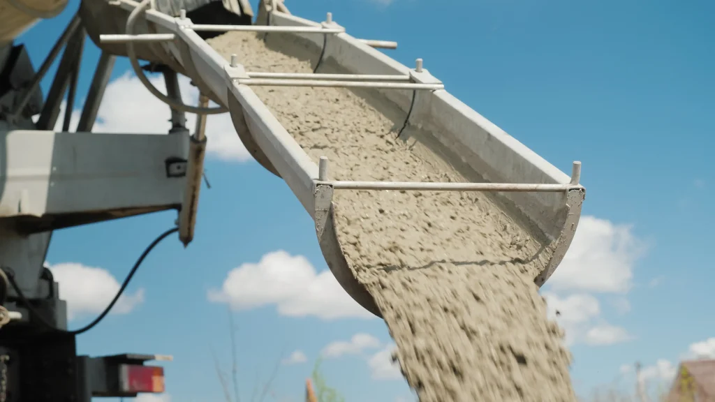 Frisch gemischter Beton fließt über eine Förderrinne aus einem Fahrmischer. Der Ortbeton wird auf einer Baustelle entladen, im Hintergrund ist blauer Himmel mit wenigen Wolken zu sehen.