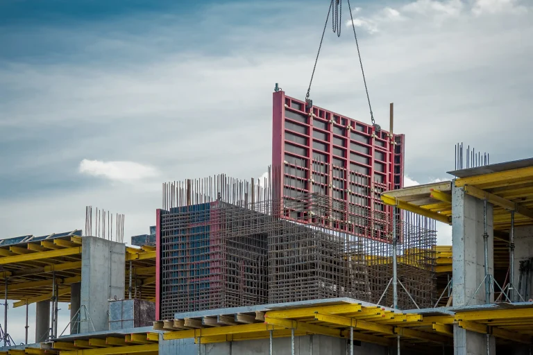 Betonschalung auf einer Baustelle im Wohnungsbau: Ein Kran hebt eine große Schalwand, während Bewehrungsstahl und Betonstützen sichtbar sind.