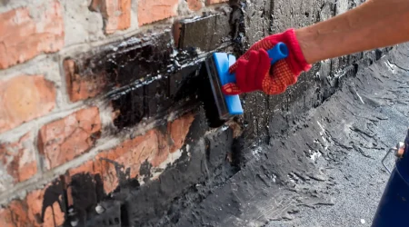 Handwerker trägt mit einer Bürste schwarze Bitumenmasse auf eine Altbau-Kellerwand aus Ziegeln auf, um das Mauerwerk gegen Feuchtigkeit abzudichten.