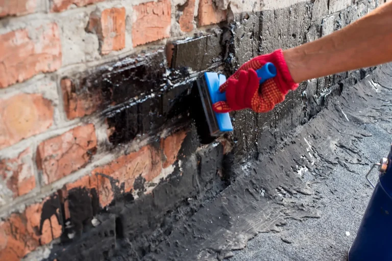 Handwerker trägt mit einer Bürste schwarze Bitumenmasse auf eine Altbau-Kellerwand aus Ziegeln auf, um das Mauerwerk gegen Feuchtigkeit abzudichten.