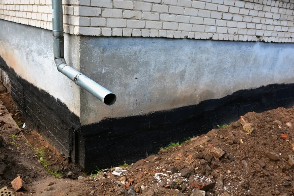 Kellerwand eines Hauses von außen mit schwarzer gestrichener Bitumenabdichtung gegen Feuchtigkeit, daneben ein Regenfallrohr aus Metall.