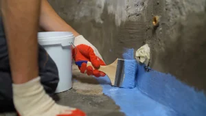 Ein Handwerker trägt mit einem Pinsel eine blaue Abdichtungsmasse an der Innenwand eines Kellers auf, um das Mauerwerk gegen Feuchtigkeit abzudichten.