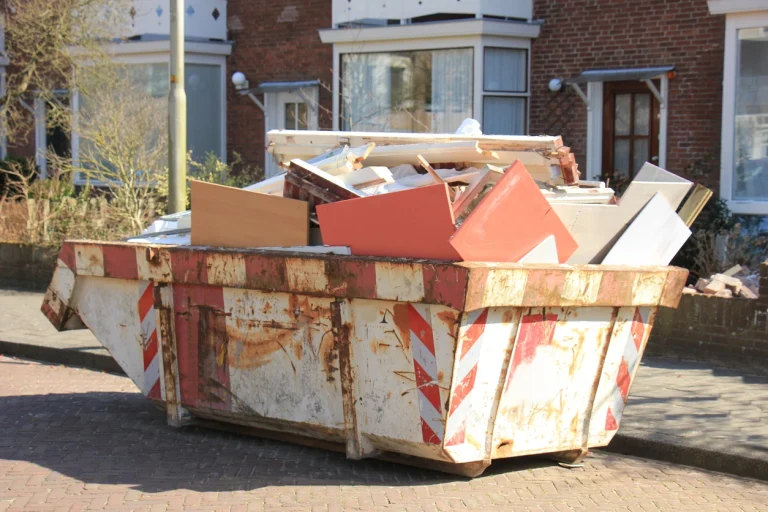 Überfüllter, rostiger Bauschuttcontainer mit Baumüll und Holz auf Bürgersteig.