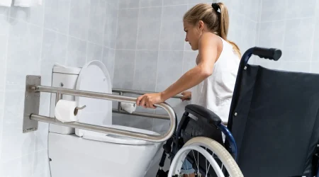 Eine Frau im Rollstuhl nutzt Haltegriffe in einem barrierefreien Badezimmer, um sich in Richtung der Toilette zu bewegen. Das barrierefreie WC ist mit stabilen Edelstahlstützgriffen und leicht zugänglichem Toilettenpapier ausgestattet.