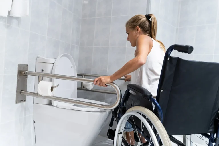 Eine Frau im Rollstuhl nutzt Haltegriffe in einem barrierefreien Badezimmer, um sich in Richtung der Toilette zu bewegen. Das barrierefreie WC ist mit stabilen Edelstahlstützgriffen und leicht zugänglichem Toilettenpapier ausgestattet.