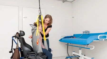Eine Pflegekraft unterstützt eine Person in einem Rollstuhl beim Transfer mit einem Deckenlifter in einem barrierefreien Badezimmer. Die Person sitzt in einer Schlinge, die an gelben Gurten befestigt ist. Im Hintergrund ist eine höhenverstellbare Pflege- oder Duschliege sichtbar