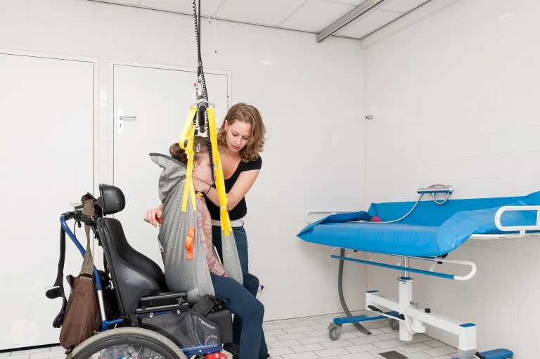 Eine Pflegekraft unterstützt eine Person in einem Rollstuhl beim Transfer mit einem Deckenlifter in einem barrierefreien Badezimmer. Die Person sitzt in einer Schlinge, die an gelben Gurten befestigt ist. Im Hintergrund ist eine höhenverstellbare Pflege- oder Duschliege sichtbar