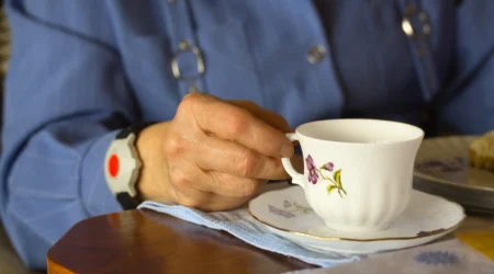 Ältere Frau mit einer dunkelblauen Bluse hält eine Porzellantasse in der Hand. An ihrem Handgelenkt trägt sie ein Notrufarmband mit GPS-Tracker mit einem großen, runden, roten Knopf, der für ihre Sicherheit sorgt.
