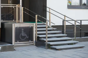 Ein barrierefreier Eingang mit einem Hublift für Rollstuhlfahrer neben einer Treppe. Der Hublift ist mit einem Symbol für Rollstuhlnutzer gekennzeichnet und verfügt über Edelstahlgeländer, die sowohl den Lift als auch die Treppe säumen.
