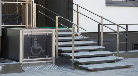 Ein barrierefreier Eingang mit einem Hublift für Rollstuhlfahrer neben einer Treppe. Der Hublift ist mit einem Symbol für Rollstuhlnutzer gekennzeichnet und verfügt über Edelstahlgeländer, die sowohl den Lift als auch die Treppe säumen.