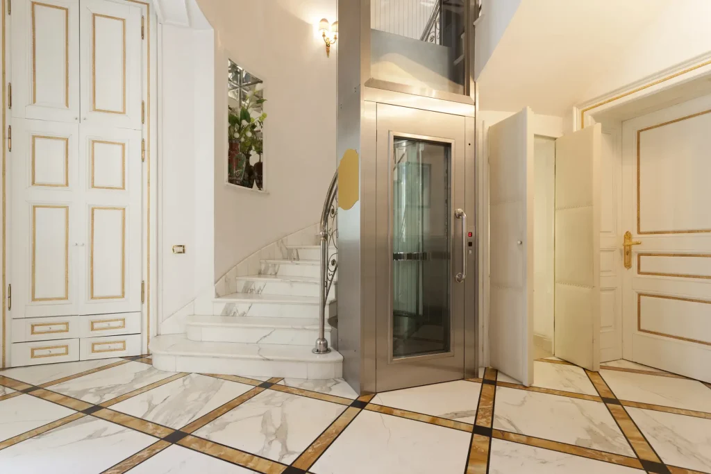 Eleganter Homelift in einem luxuriösen Wohnhaus mit Marmorböden und einer geschwungenen Treppe. Der gläserne Aufzug mit Edelstahlrahmen fügt sich nahtlos in die klassische, stilvolle Innenarchitektur ein. Der Lift ist neben einer offenen Tür platziert und ermöglicht eine barrierefreie Verbindung zwischen den Stockwerken.