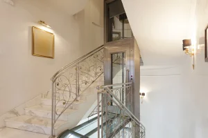 Homelift in einem eleganten Treppenhaus mit kunstvoll verzierten Geländern und Marmorstufen. Der Aufzug ist in einem modernen Glas-Edelstahl-Design gestaltet und bietet eine barrierefreie Lösung für die Verbindung der Stockwerke. Die edlen Wandlampen und ein goldgerahmter Spiegel sorgen für eine luxuriöse Atmosphäre.