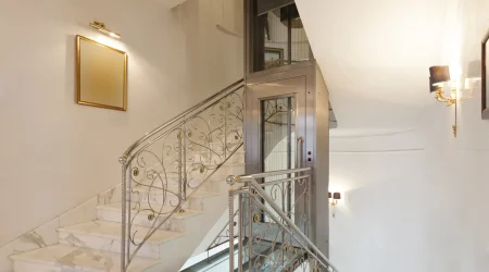 Homelift in einem eleganten Treppenhaus mit kunstvoll verzierten Geländern und Marmorstufen. Der Aufzug ist in einem modernen Glas-Edelstahl-Design gestaltet und bietet eine barrierefreie Lösung für die Verbindung der Stockwerke. Die edlen Wandlampen und ein goldgerahmter Spiegel sorgen für eine luxuriöse Atmosphäre.