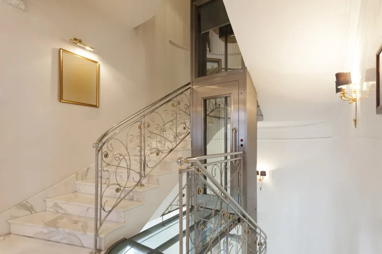 Homelift in einem eleganten Treppenhaus mit kunstvoll verzierten Geländern und Marmorstufen. Der Aufzug ist in einem modernen Glas-Edelstahl-Design gestaltet und bietet eine barrierefreie Lösung für die Verbindung der Stockwerke. Die edlen Wandlampen und ein goldgerahmter Spiegel sorgen für eine luxuriöse Atmosphäre.
