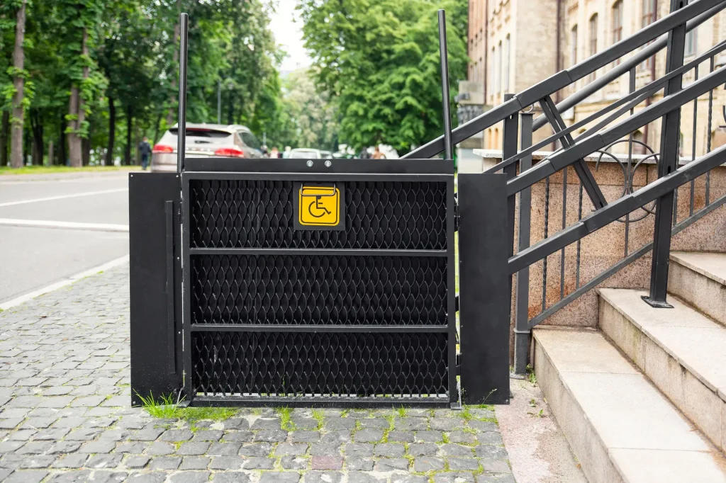 Ein barrierefreier Hebelift mit einem schwarzen Metallgitterdesign neben einer Treppe. Der Lift ist mit einem gelben Symbol für Rollstuhlfahrer gekennzeichnet und steht auf einem gepflasterten Gehweg.