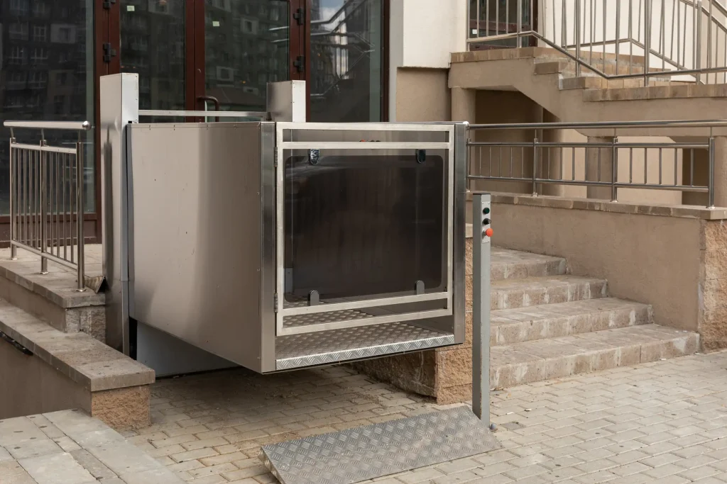 Hublift für Rollstuhlfahrer an einem Eingang mit Treppenstufen. Der Hebelift ist aus Metall und verfügt über ein transparentes Sicherheitsgeländer. Eine Rampe erleichtert den Zugang zur Plattform, die durch Bedienelemente an einem externen Bedienpult gesteuert wird. Der Hublift ist ideal für den barrierefreien Zugang zu Gebäuden in städtischen Umgebungen und ermöglicht Menschen mit eingeschränkter Mobilität eine selbstständige Nutzung.