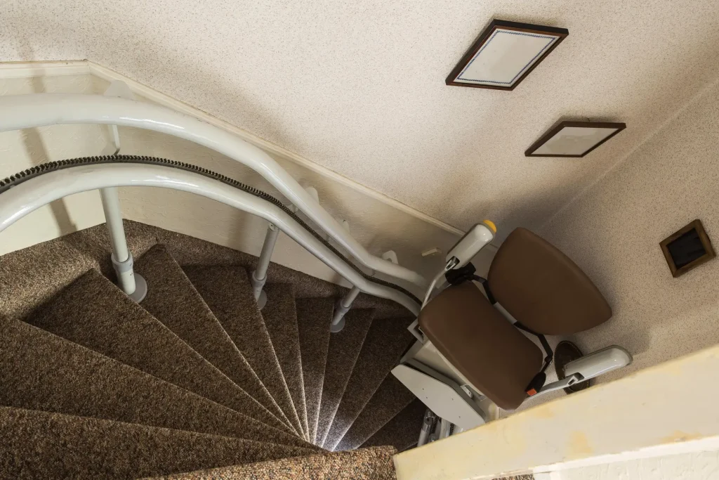 Kurvenlift auf einer engen, gewundenen Treppe in einem Wohngebäude. Der Treppenlift ist an doppelten Schienen montiert und folgt dem Verlauf der Wendeltreppe, um Menschen mit eingeschränkter Mobilität den Zugang zu ermöglichen. Die Treppe ist mit Teppichboden versehen, und der Lift verfügt über einen gepolsterten Sitz und Armlehnen für zusätzlichen Komfort.