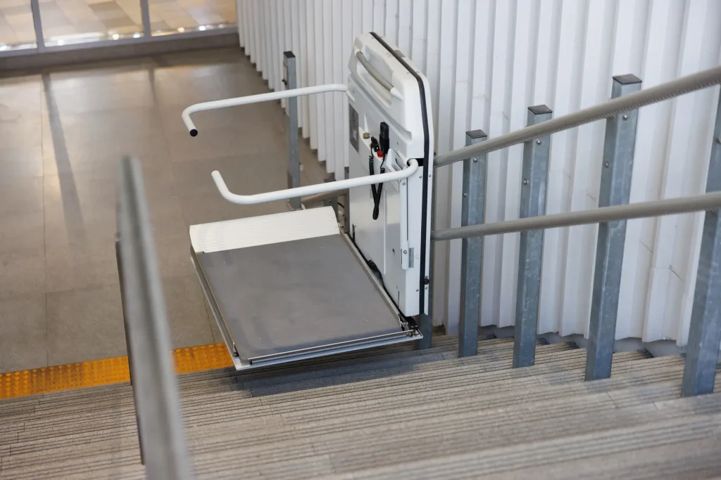 Rollstuhllift auf einer Treppe in einem öffentlichen Gebäude. Der Plattformlift ist mit Haltegriffen und einer breiten Fläche ausgestattet, um Rollstuhlfahrern den Zugang zu ermöglichen. Die Schienen verlaufen parallel zur Treppe und die Sicherheitsvorrichtungen sind sichtbar.