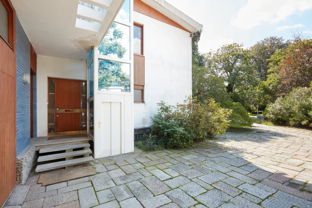 "Senkrechtlift an der Außenseite eines modernen Wohnhauses mit direktem Zugang zur Eingangstür. Der Aufzug ist in einem weißen, geschlossenen Schacht integriert und ermöglicht barrierefreien Zugang zu höheren Ebenen. Die Umgebung ist von gepflasterten Wegen, üppigem Grün und Bäumen umgeben. Das Haus hat eine helle Fassade mit hölzernen Akzenten, die dem Gebäude ein stilvolles Aussehen verleihen.