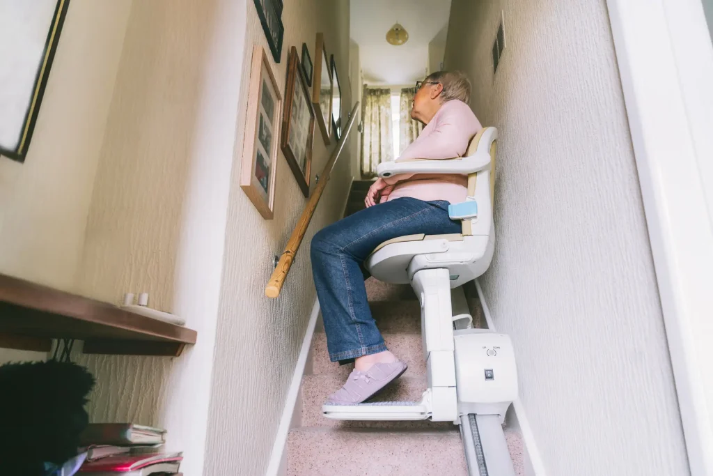 Ältere Dame sitzt auf einem Treppenlift in einem schmalen Treppenhaus eines Wohnhauses. Der Sitzlift ist auf einer Schiene an der Treppenwand installiert und ermöglicht der Frau eine sichere und bequeme Beförderung nach oben. Die Frau trägt eine Brille, ein rosa Oberteil und Jeans, während sie entspannt auf dem Lift sitzt. An der Wand sind Bilderrahmen und ein Handlauf zu sehen.