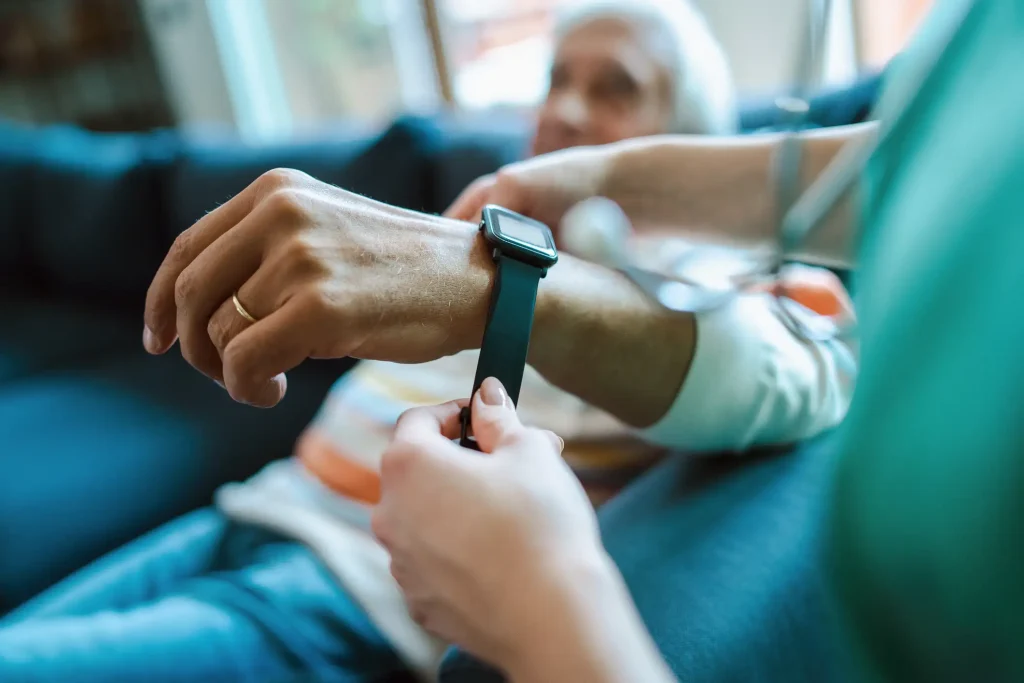 Pflegekraft befestigt eine Smartwatch mit Sturzsensor am Handgelenk eines älteren Menschen. Die Smartwatch ist schwarz und bietet moderne Sicherheitsfunktionen, die speziell für Senioren entwickelt wurden. Im Hintergrund ist der Senior unscharf zu erkennen, während er entspannt auf einem Sofa sitzt.