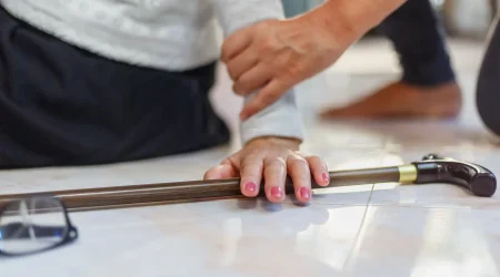 Eine Seniorin liegt nach einem Sturz auf dem Boden, während eine helfende Hand sie stützt, um aufzustehen. Auf dem Boden liegen ein Gehstock und eine Brille.