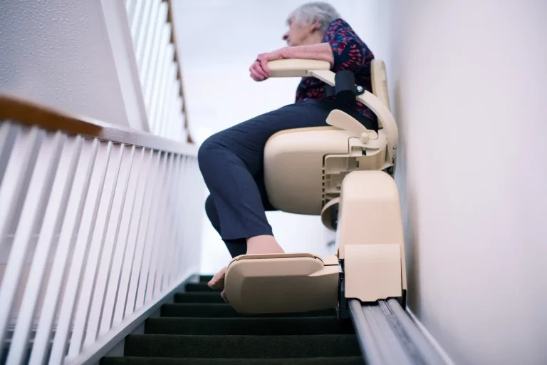 Seniorin sitzt auf einem modernen Treppenlift, der entlang einer Schiene an einer Teppichtreppe montiert ist. Die Frau lehnt sich entspannt auf die Armlehne, während der Lift sie sicher nach oben transportiert. Sie trägt eine gemusterte Bluse und dunkle Hosen.