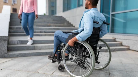 Ein Mann im Rollstuhl betrachtet eine Treppe vor einem modernen Gebäude, während eine Frau die Treppe hinuntergeht. Das Bild verdeutlicht die Herausforderungen für barrierefreies Bauen im öffentlichen Raum.
