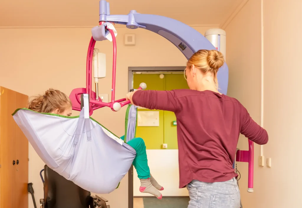 Pflegekraft nutzt einen mobilen Hängelifter, um ein Kind sicher in eine Sitzposition zu bringen.