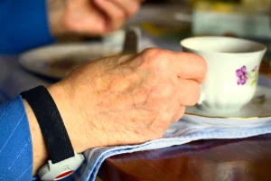 Eine Nahaufnahme einer älteren Hand, die eine Tasse hält, während am Handgelenk ein mobiler Notrufknopf getragen wird.