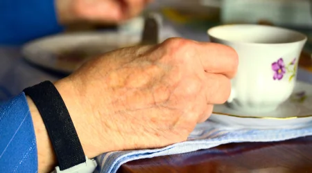 Eine Nahaufnahme einer älteren Hand, die eine Tasse hält, während am Handgelenk ein mobiler Notrufknopf getragen wird.