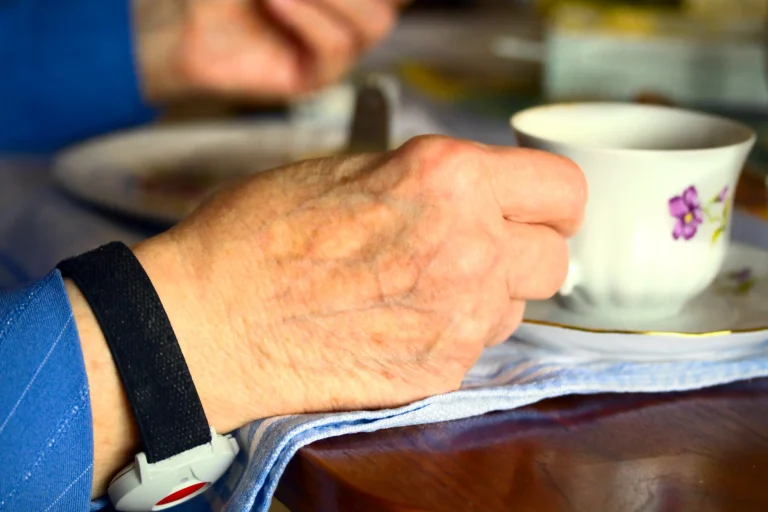 Eine Nahaufnahme einer älteren Hand, die eine Tasse hält, während am Handgelenk ein mobiler Notrufknopf getragen wird.